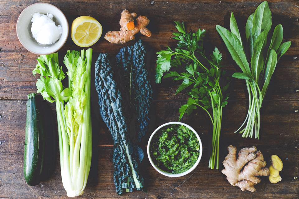 Overhead view of Green Juju Just Greens Frozen Blend ingredients: coconut oil, lemon, turmeric, zucchini, celery, kale, herb purée, parsley, leafy greens, ginger.
