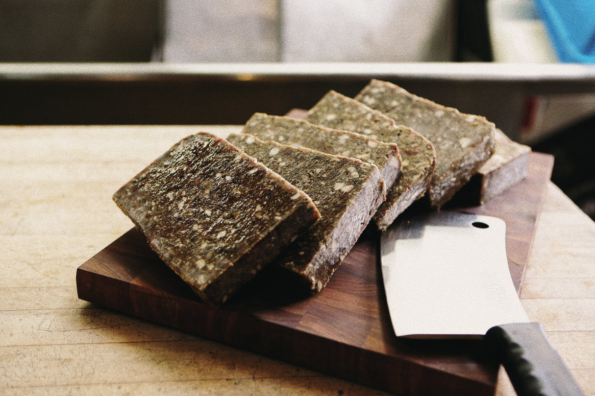 Butcher Blend Beef, Salmon & Tripe for Dogs; thick, gray-brown meat loaf slices with fat flecks on a wooden board, cleaver beside.