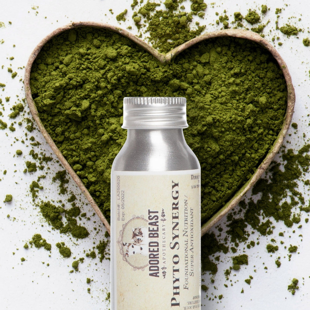 Adored Beast Phyto Synergy supplement bottle beside a heart-shaped dish filled with bright green powder, scattered on a white background.