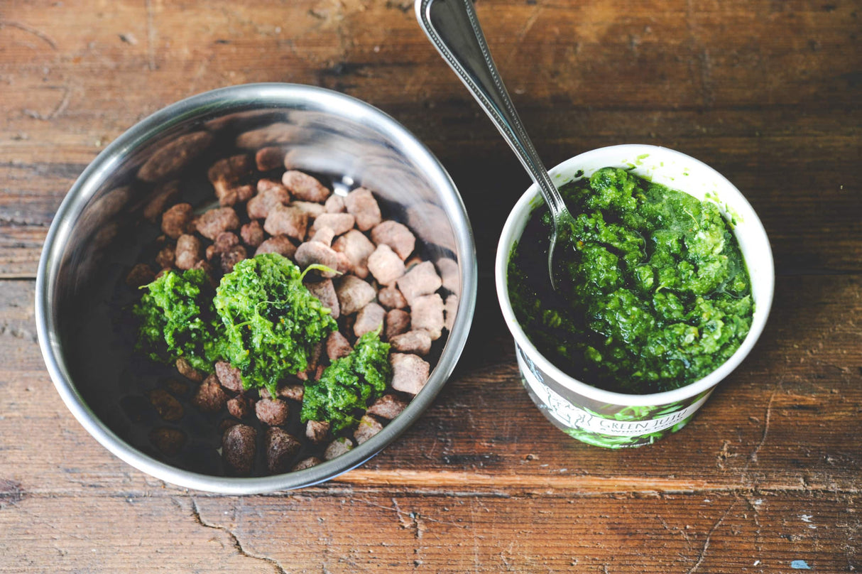 Green Juju Just Greens Frozen Blend 15oz in a stainless bowl atop kibble, with tub of green vegetable puree and spoon beside it on wooden table.