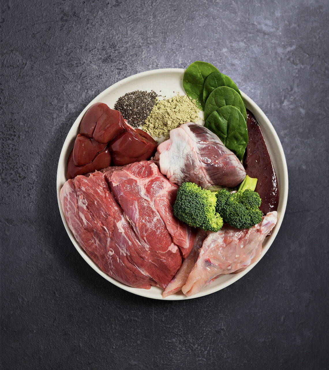 Rawbone Mixed Protein Lamb Meal platter with raw lamb roast, chicken pieces, organ meats, broccoli, spinach, seeds, and powders, ideal for homemade pet diets.