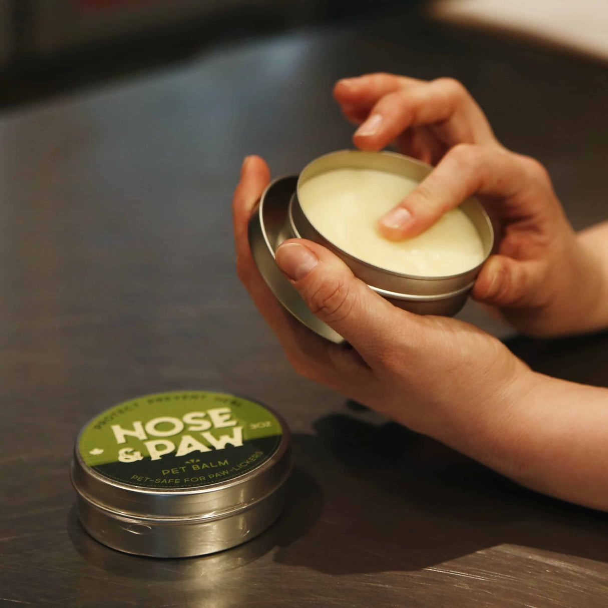 Carnivore Care Nose & Paw Balm tins on a table, one open with a fingertip in the balm, highlighting pet-safe, soothing, natural ingredients.