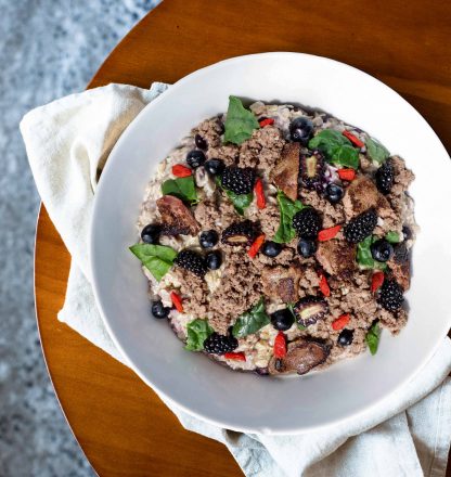 Tom & Sawyer Goji Berry Beef: a bowl of cooked grains topped with seasoned beef, fresh berries, greens, goji berries, and thin dark slices on a cloth napkin.