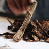 Hand reaches for dark-brown One Ingredient Dehydrated Beef Brisket jerky strips on white surface, featuring True Carnivores watermark, ideal for pet training or rewarding.