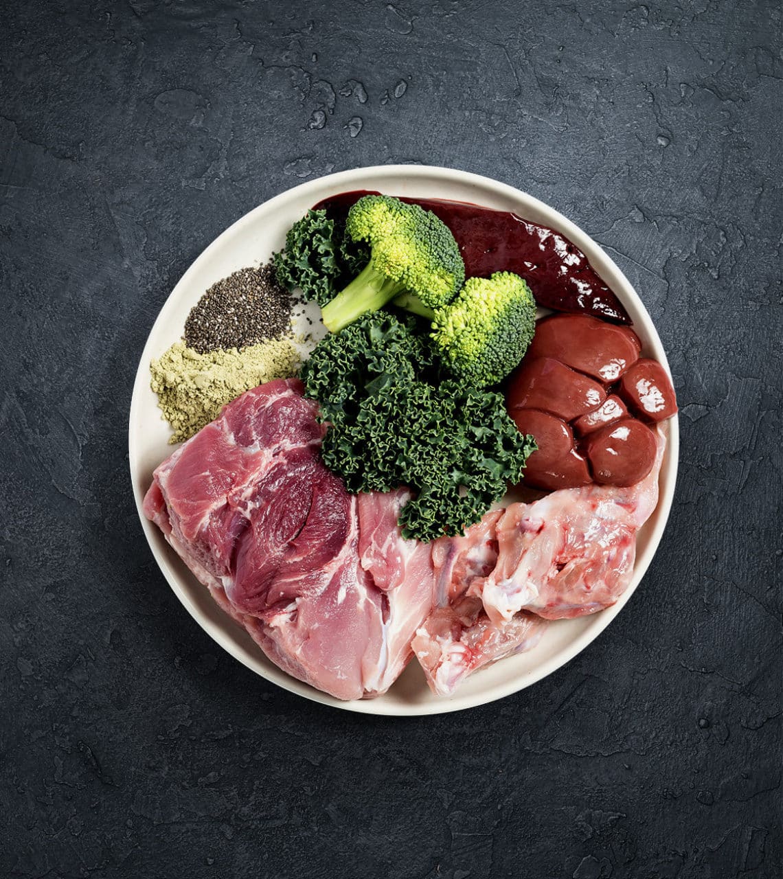 Rawbone Mixed Protein Pork Meal in a bowl: raw pork, organ meats, bone, kale, broccoli, chia seeds, green powder, and red puree. Ideal for raw diets.