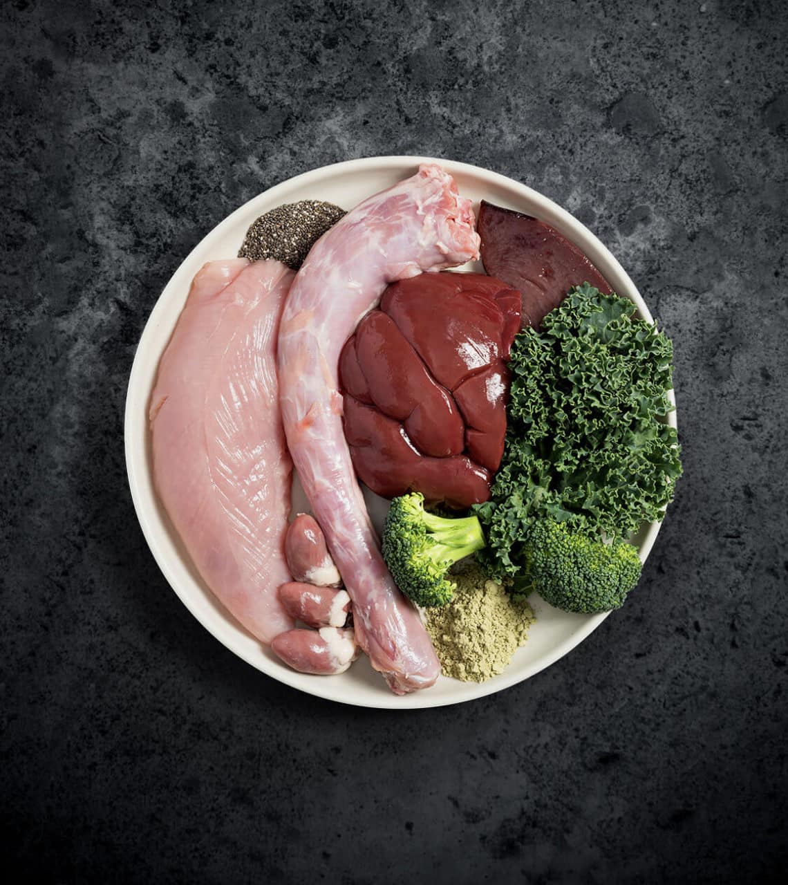 Rawbone Mixed Protein Turkey Meal in a white bowl with raw ingredients: turkey neck, liver, kale, broccoli, chia seeds, green powder, organs, and muscle-meat filet.